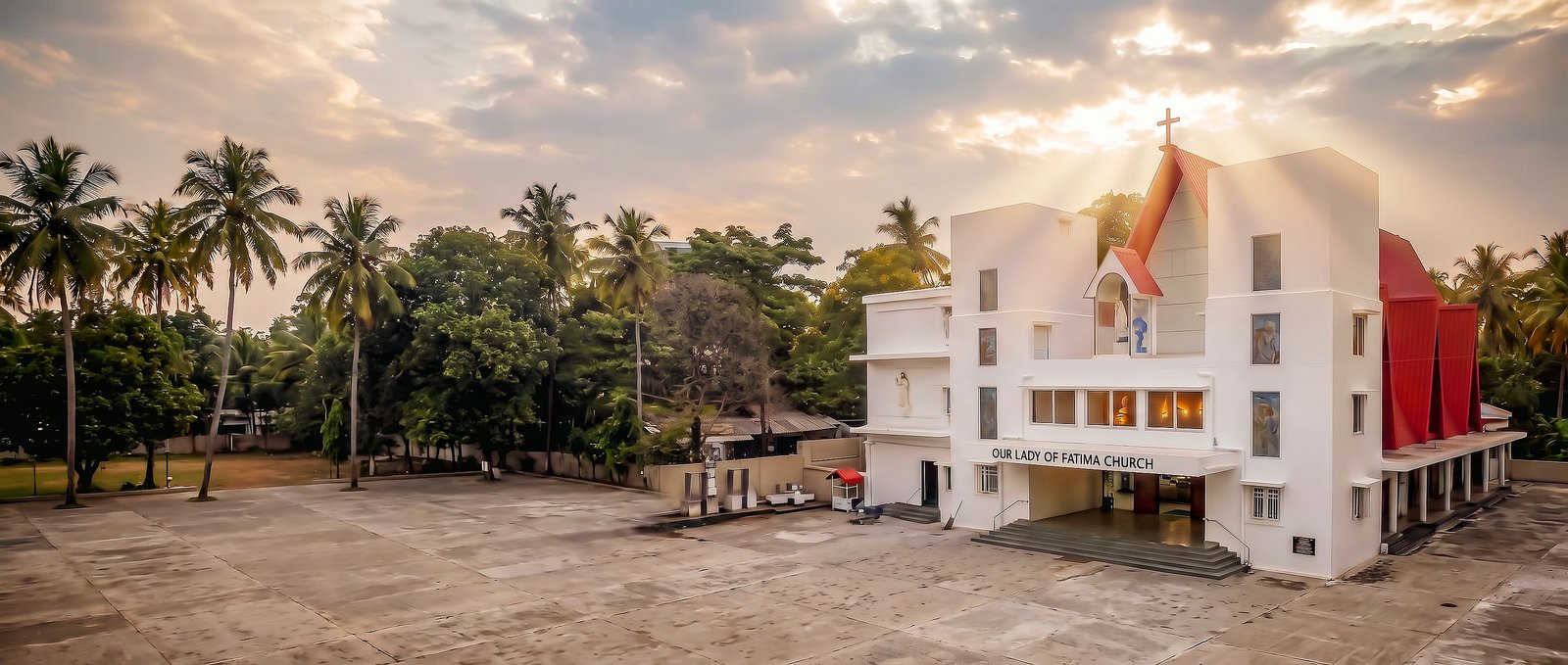 Our Lady of Fatima Church at golden hour across its open forecourt: the white façade, the red roof and the cross, with sunlight breaking through the clouds behind it and coconut palms around the compound