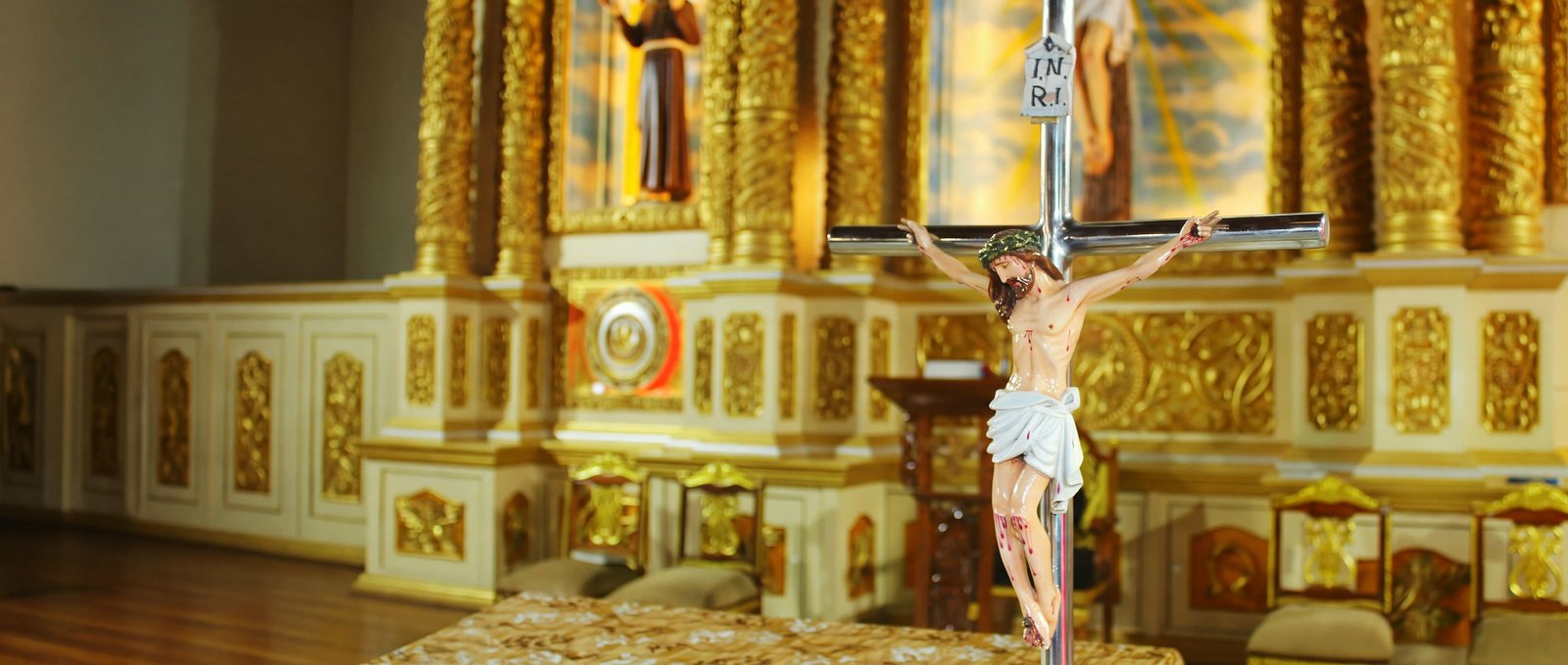 The altar crucifix of Our Lady of Fatima Church standing before the gilded reredos, with the sanctuary and its carved altar rail receding into the quiet of the church beyond