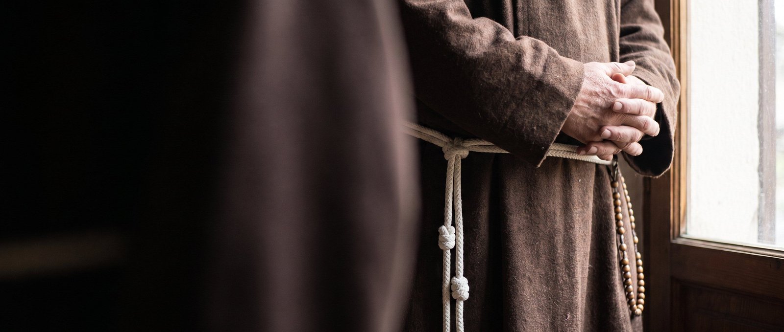 A friar’s hands folded over his brown habit, with its knotted white cord and a wooden rosary